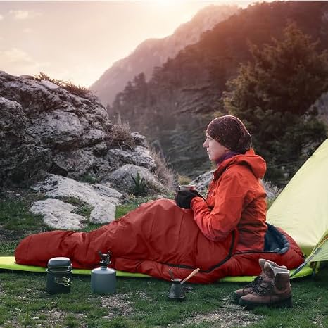 A person sits in a red sleeping bag on a grassy mountain camping site at sunset, with a yellow tent, rocks, trees, and camping gear visible.