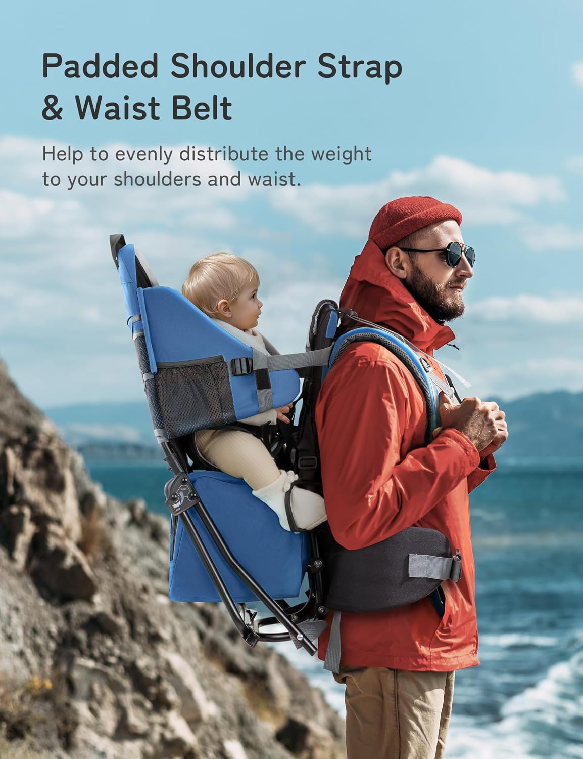 Man carrying a child in a blue chair with a padded shoulder strap and waist belt, standing on a rocky cliff overlooking the ocean.