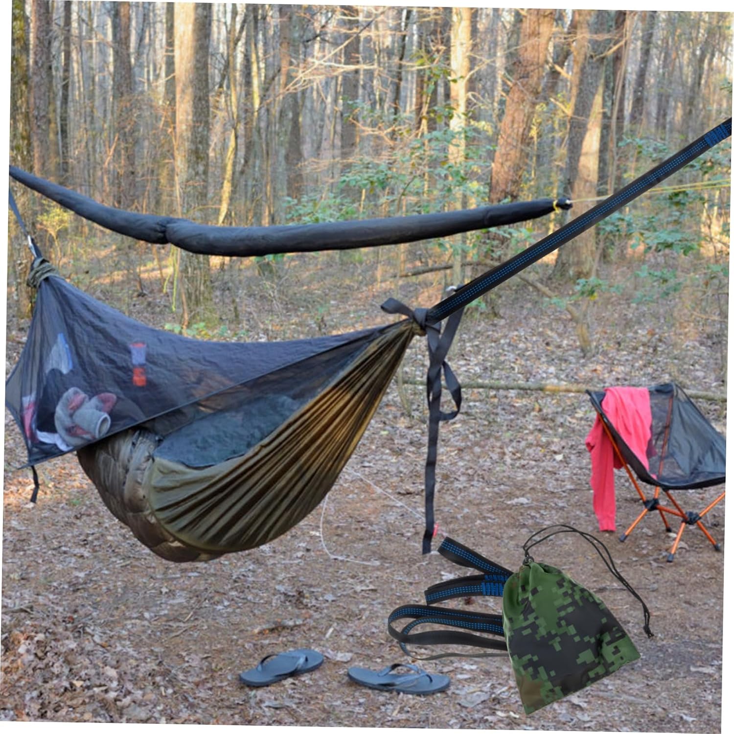 A camping hammock set up in a wooded outdoor area, suspended between two black ground anchors with a dark mesh fly sheet. A sleeping pad is visible inside the hammock, with a camouflage drawstring bag, straps, and a pink garment hanging on a folding chair nearby.
