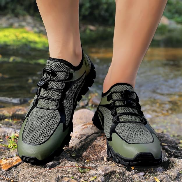 A pair of men's athletic hiking shoes in olive green and black, shown being worn on a person's feet standing on rocks near a stream.