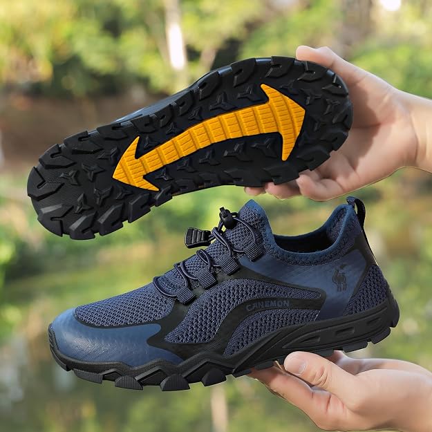 A pair of dark blue and black athletic trail shoes being held by hands to show both the top and the treaded sole. The sole features a prominent yellow arrow graphic indicating direction.