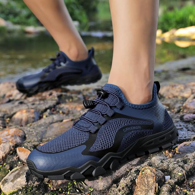 A person wearing dark blue and black outdoor slip-on hiking shoes standing on rocks near a stream, with the second shoe visible in the background.