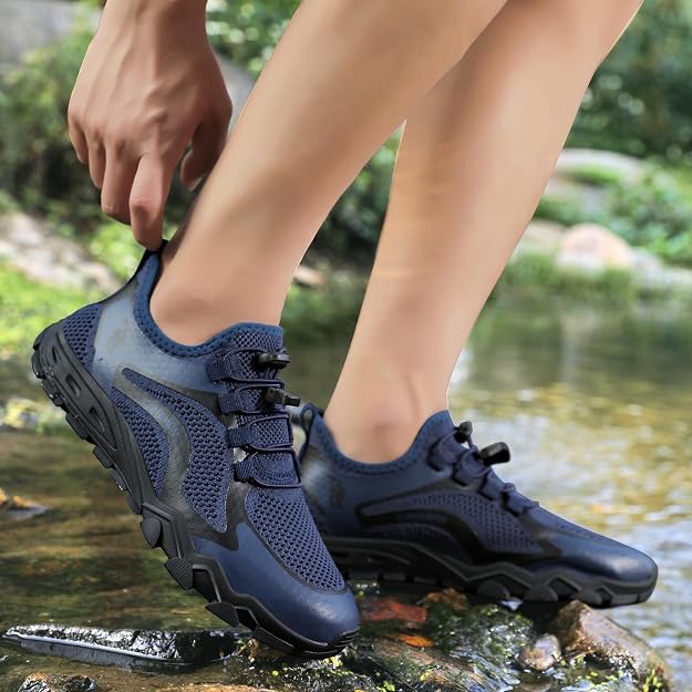 A person wearing dark blue water-resistant hiking shoes, with one shoe being adjusted by a hand, standing on a rock in a shallow stream with a blurred natural background. big green bear