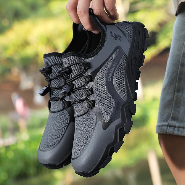 A pair of dark gray and black athletic hiking shoes being held by a person's hand, with a blurred outdoor green background and part of the person's denim shorts visible on the right.