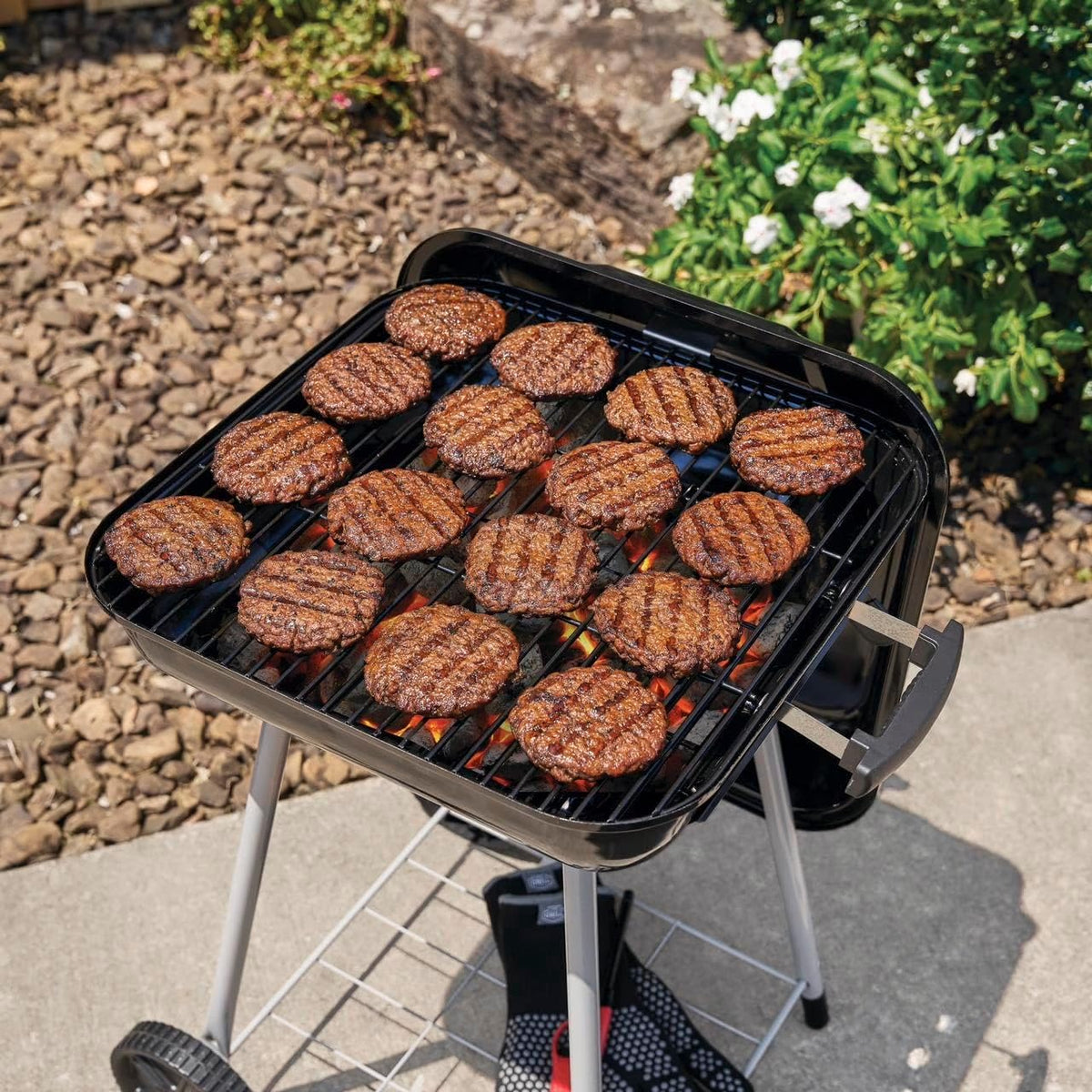 A black charcoal grill with silver legs outdoors on a patio, cooking multiple grilled patties over visible coals. Rocks, greenery, and flowers are in the background.