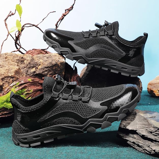 A pair of black athletic trail running shoes with mesh and synthetic overlays, displayed on rocks against a blue background.
