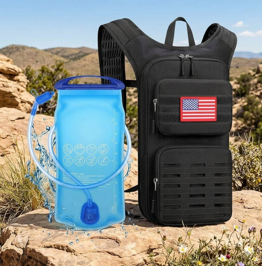A black hydration backpack shown outdoors on a rocky desert landscape next to a translucent blue water bottle with a drinking tube. The backpack has multiple zippered compartments, a top handle, shoulder straps, and a small American flag patch on the front pocket.