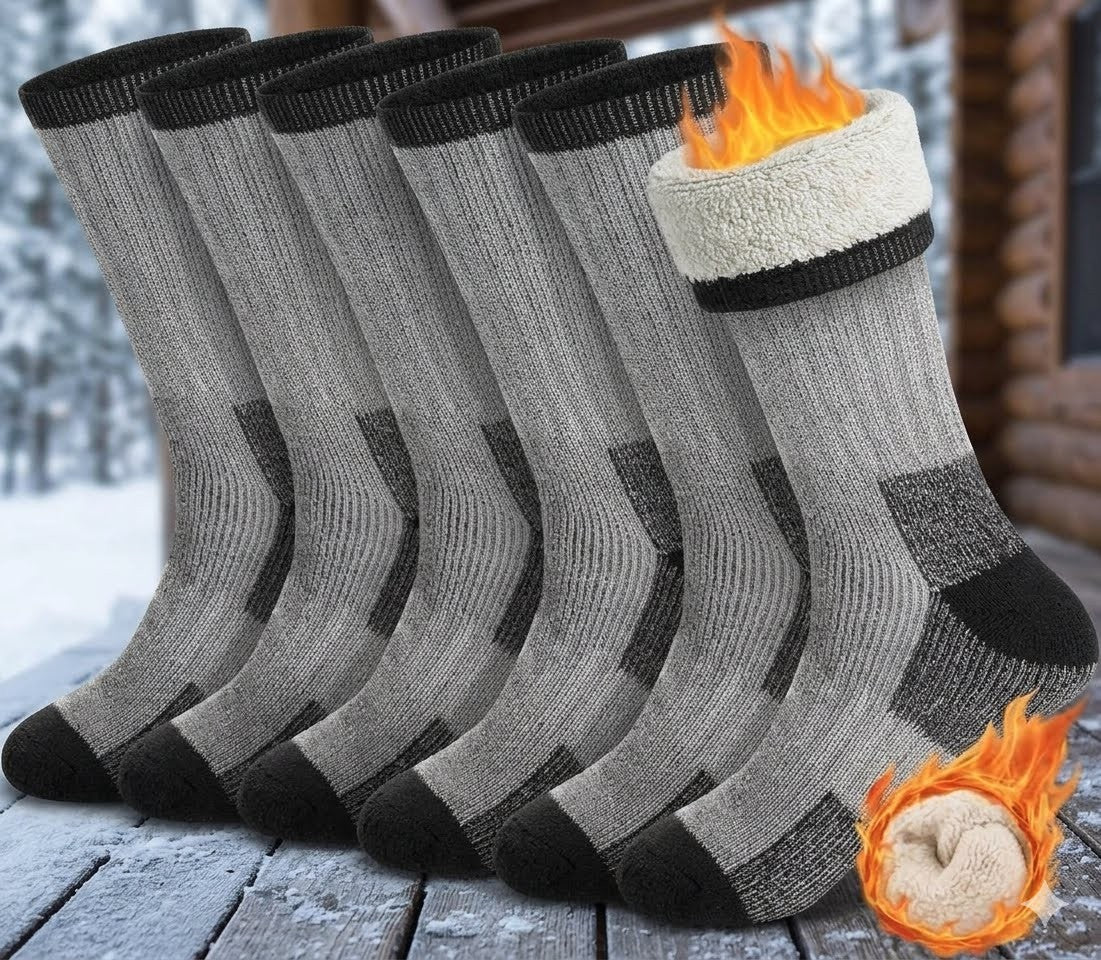 Five gray knit socks with black heels, toes, and cuffs are shown on a snowy wooden deck. One sock is lifted to reveal a white fleece lining with a flame graphic, and a second flame graphic appears in the lower right over a beige inner lining. big green bear