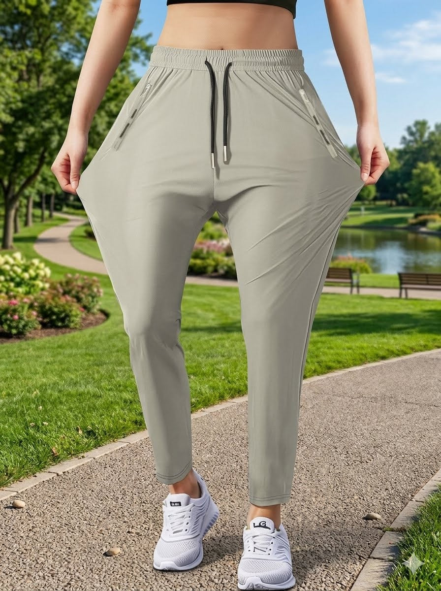 A person outdoors on a park path wearing loose, high-waisted athletic jogger pants in a light sage green color, holding the sides of the pants to show their stretch. The background shows a green park with grass, trees, and a pond.