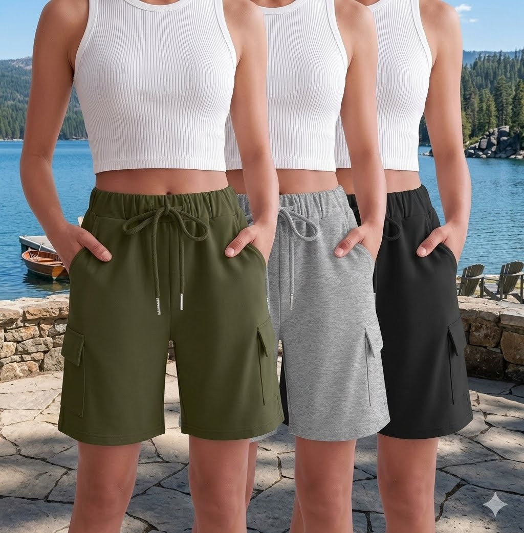 Three women shown from the chest down wearing matching cargo shorts in olive green, heather gray, and black, standing outdoors by a lake. big green bear
