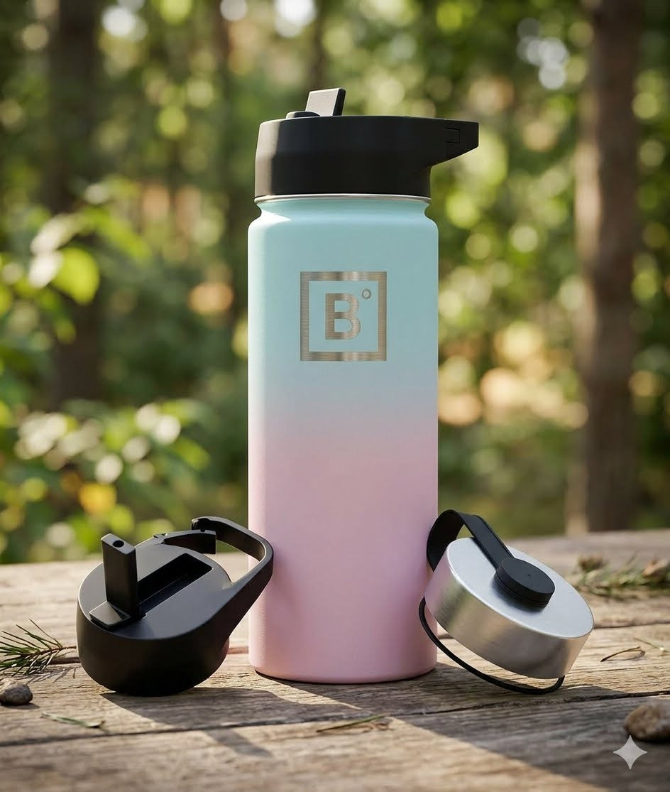 A reusable insulated water bottle with a black flip-straw lid, shown standing on a wooden surface outdoors with a blurred forest background. The bottle has a pastel gradient from light aqua at the top to soft pink at the bottom, with a metallic square logo featuring a stylized letter B on the front. big green bear
