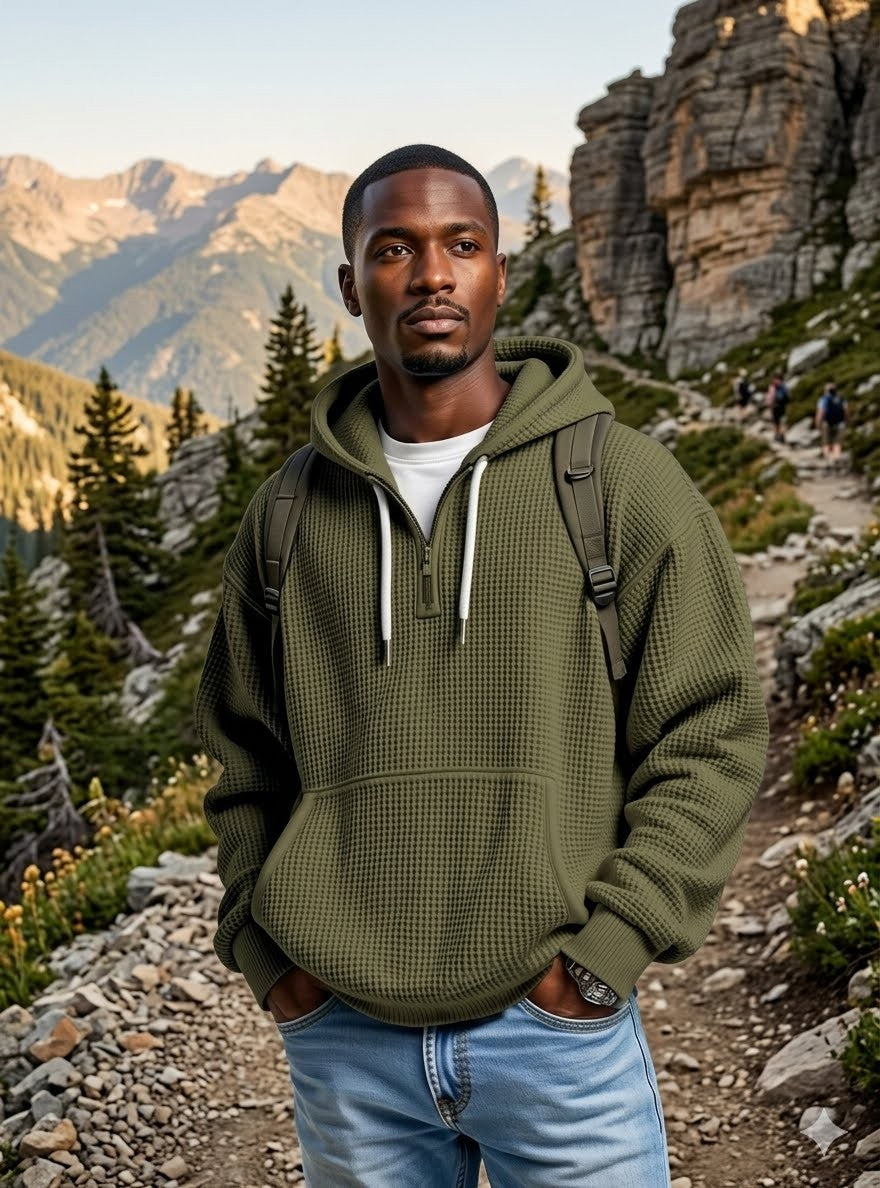 A man wearing an olive green waffle-knit pullover hoodie with white drawstrings, standing on a rocky mountain trail with pine trees and cliffs in the background. big green bear