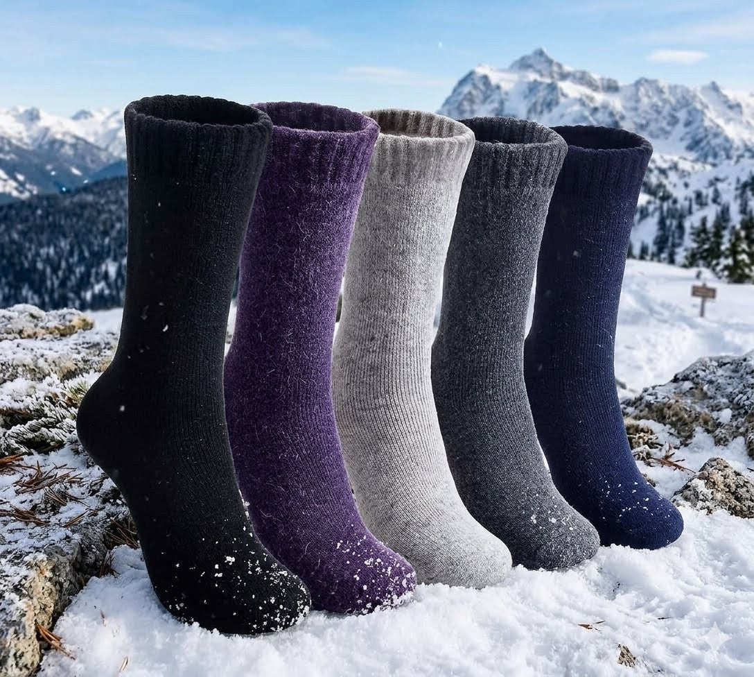 Five pairs of mid-calf knit socks in black, purple, light gray, dark gray, and navy blue, arranged upright in the snow with a snowy mountain landscape in the background. big green bear