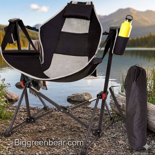 A black and gray folding camp chair with a side cup holder holding a yellow water bottle, set outdoors by a lake with mountains in the background. A black carrying bag stands beside the chair, and a Biggreenbear.
