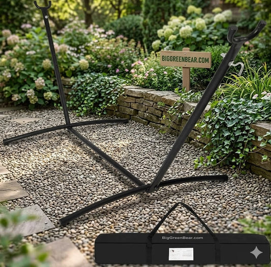 A black metal outdoor swing stand displayed on a gravel path in a landscaped garden, with its carrying case shown in the foreground and a sign reading BIGGREENBEAR.COM in the background.