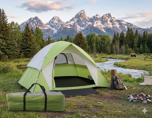 A green and white dome camping tent is pitched in a grassy clearing with a stream, pine trees, and snow-capped mountains in the background. A matching green tent bag sits in the foreground, with a backpack, chair, and fire pit nearby. big green bear biggreenbear.com