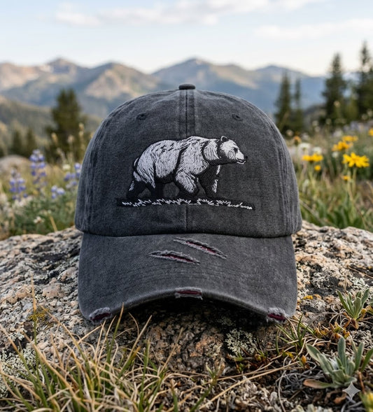A dark gray distressed baseball cap with an embroidered black and white grizzly bear design, resting on a large rock in a mountain meadow with wildflowers and distant mountains in the background. big green bear biggreenbear.com