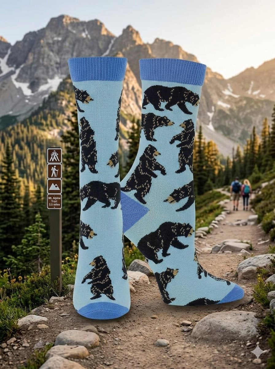 A pair of light blue crew socks with a black and brown bear print, displayed on a hiking trail with mountains and forest in the background. big green bear biggreenbear.com