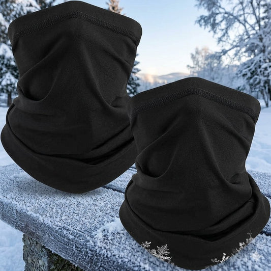 Two black fleece face masks or neck gaiters displayed outdoors on a snow-covered stone ledge, with a snowy forest background. big green bear biggreenbear.com