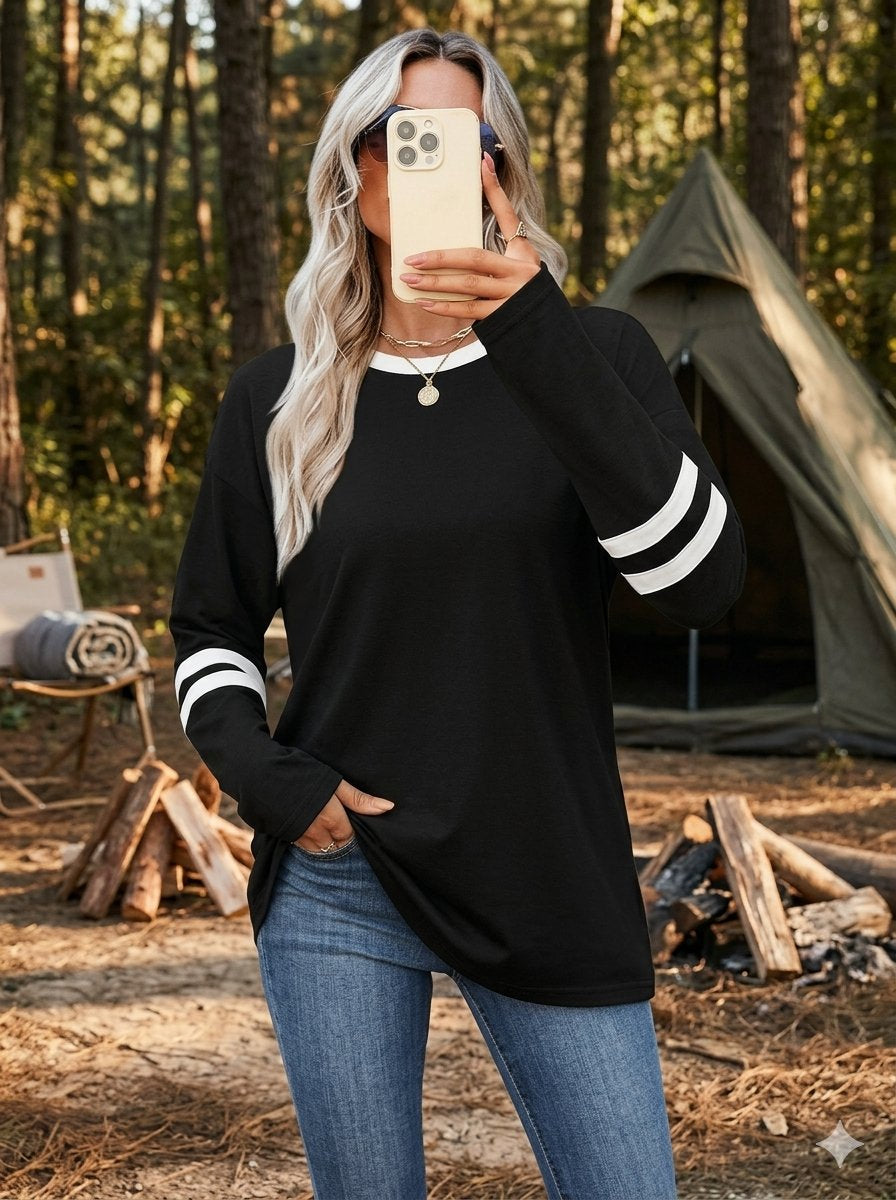 A woman takes a mirror selfie in a forest campsite setting, wearing a black long-sleeve t-shirt with white stripes on the cuffs and blue jeans. big green bear biggreenbear.com