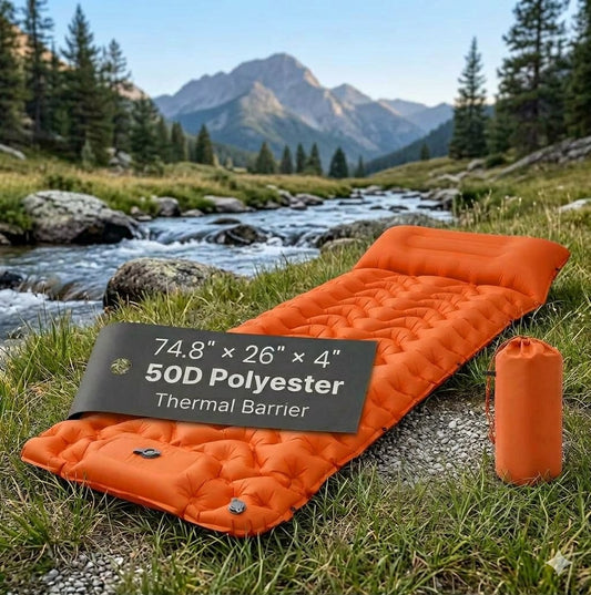 An orange inflatable camping sleeping pad laid out on grass beside a stream, with a mountain landscape in the background. A black label on the pad reads '74. big green bear biggreenbear.com