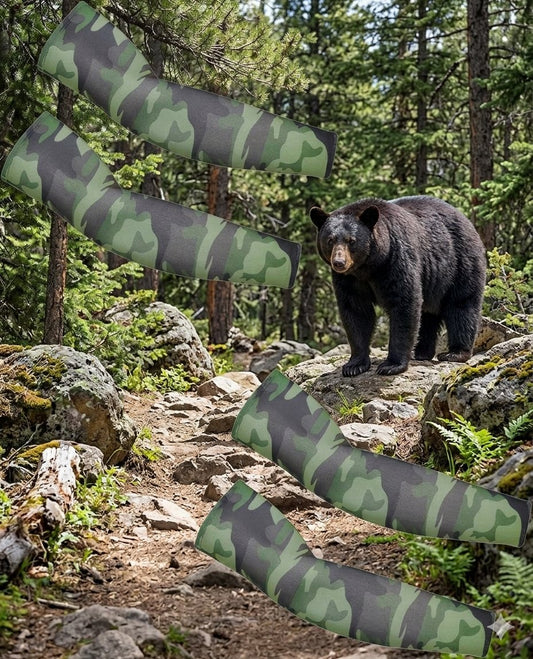 Four camouflage-patterned arm warmers are shown floating over a forest trail with a black bear standing on rocks in the background. The camouflage design features shades of green, gray, and black. big green bear biggreenbear.com