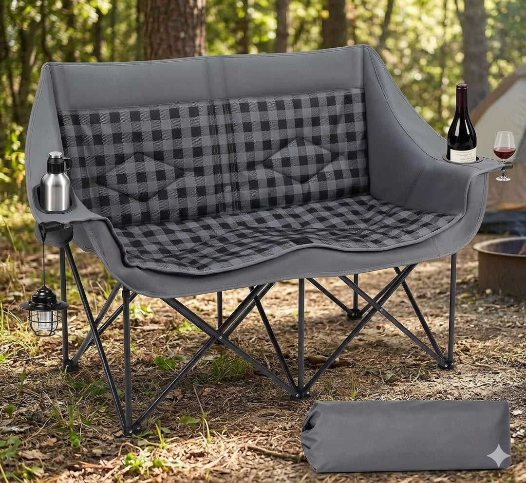 A gray folding camp chair with a black-and-gray buffalo plaid seat cushion, shown outdoors in a wooded campsite setting with a bottle, wine glass, lantern, and a matching gray carry bag on the ground. big green bear biggreenbear.com