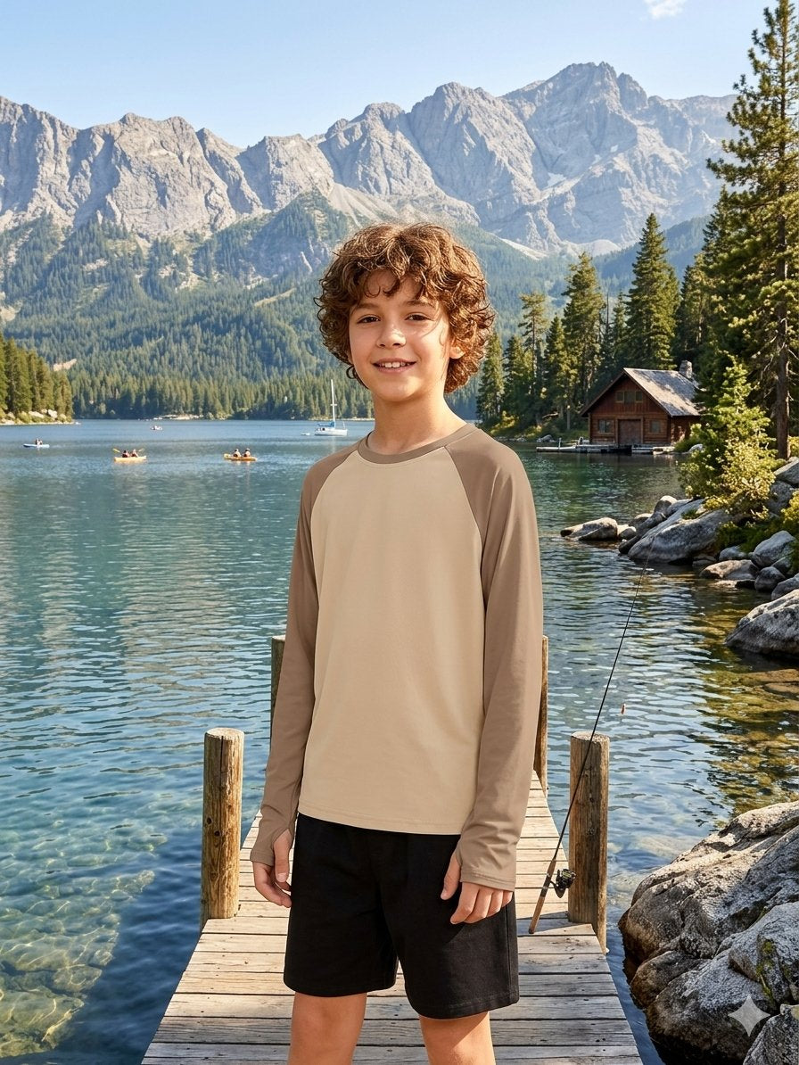 A young boy stands on a wooden dock by a calm lake with mountains, trees, and a small cabin in the background. He is wearing a long-sleeve athletic shirt with a beige torso and brown raglan sleeves, paired with black shorts. big green bear biggreenbear.com
