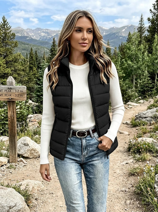 A woman standing outdoors on a mountain trail wearing a black puffer vest over a white ribbed long-sleeve top and blue jeans, with mountains and trees in the background. big green bear