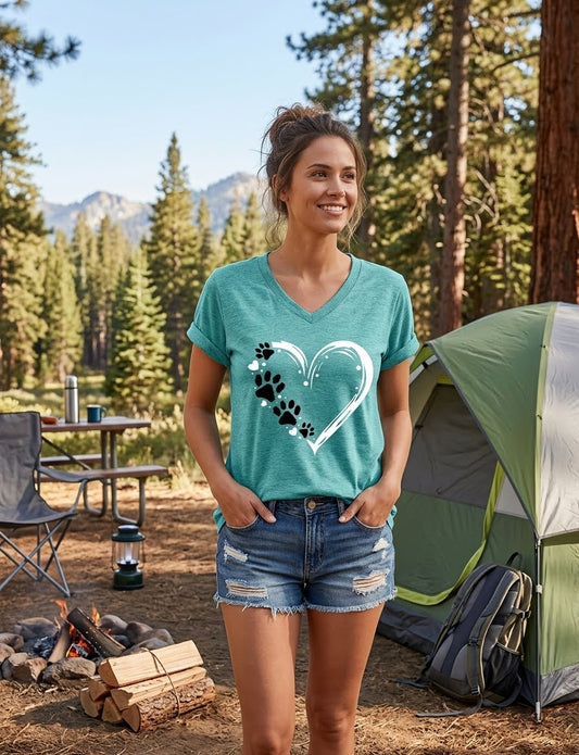 A woman stands outdoors in a campsite setting wearing a teal heathered V-neck t-shirt with a white heart outline and black paw print graphic, paired with denim shorts. A green tent, campfire, and pine trees are visible in the background. big green bear