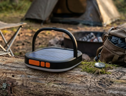 A portable solar lantern with a built-in handle sits on a weathered log in an outdoor campsite setting, with a tent, backpack, and fire pit visible in the background. big green bear biggreenbear.com