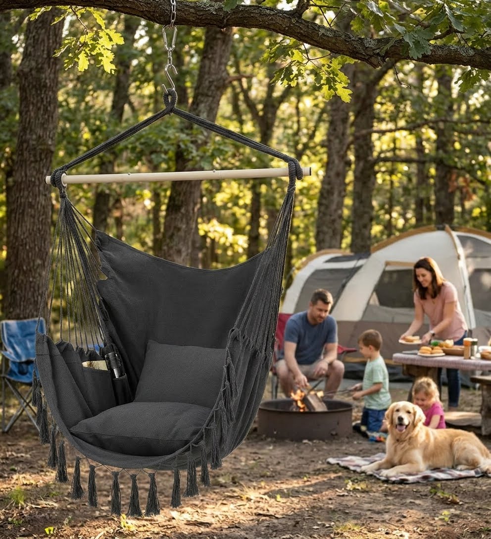 A dark gray fabric hammock swing with tassels hangs from a tree at a forest campsite, with a family, a dog, and a tent in the background.