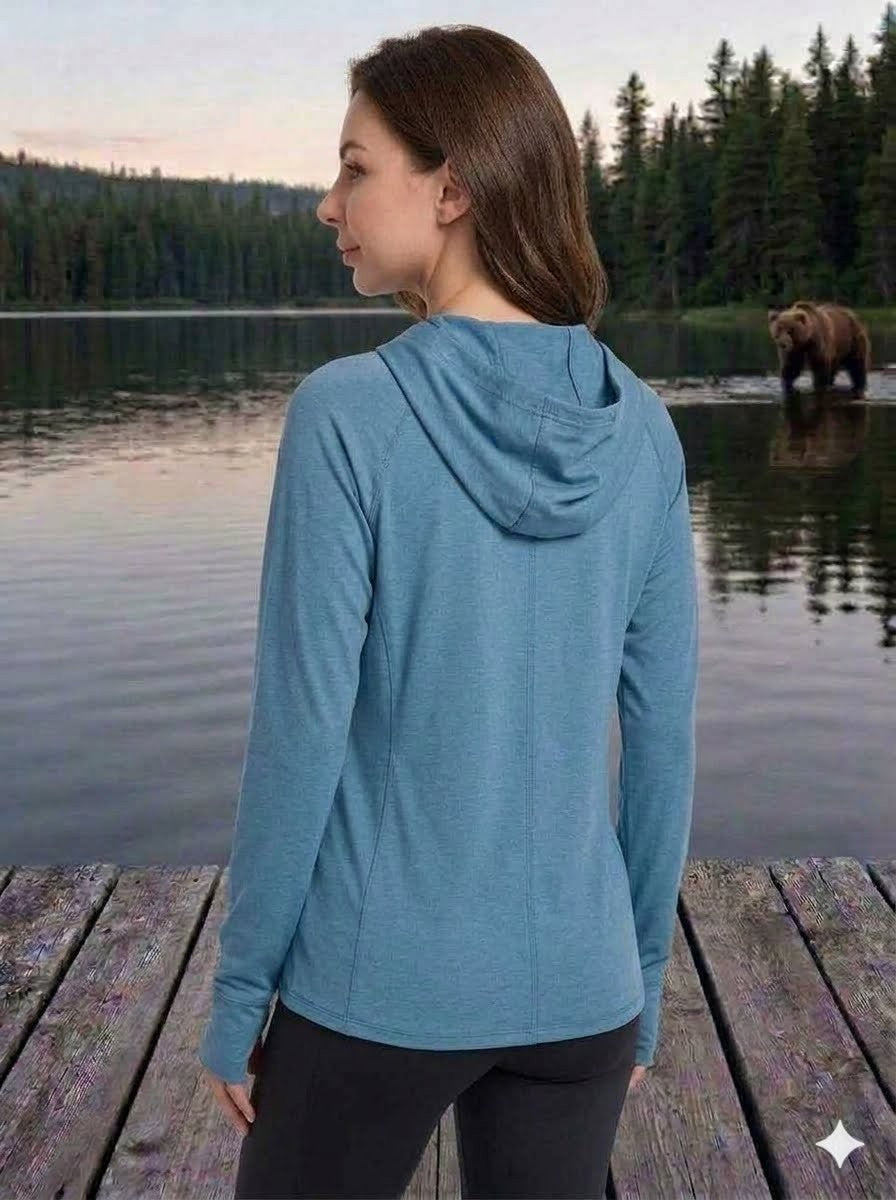 Woman in a blue hoodie standing on a wooden dock by a lake with a bear in the background.