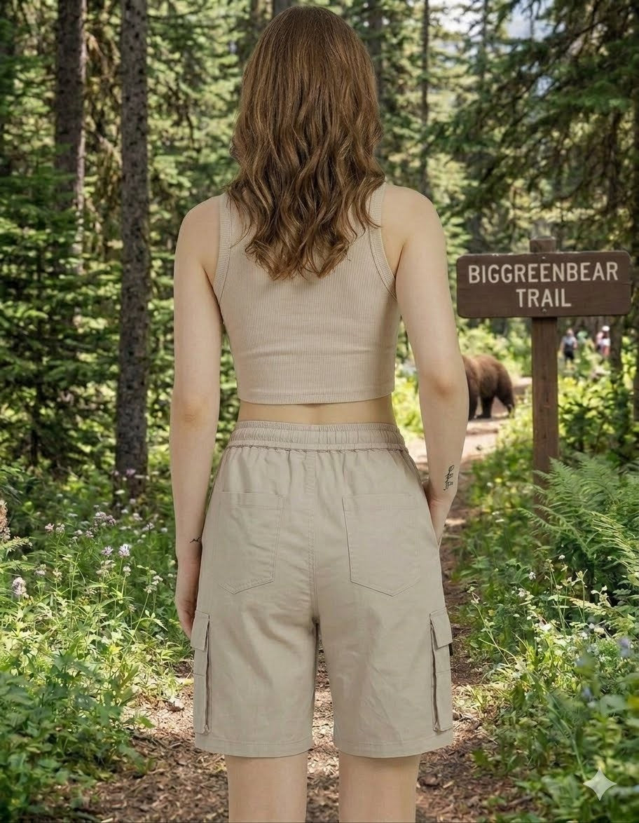 Person in beige top and shorts standing on a trail with a 'Biggreenbear Trail' sign in a forest.