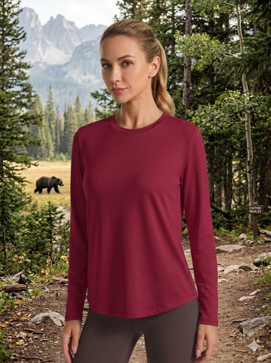 A woman wearing a long-sleeve crewneck athletic top in burgundy, standing in a forest with mountains and a bear in the background. big green bear