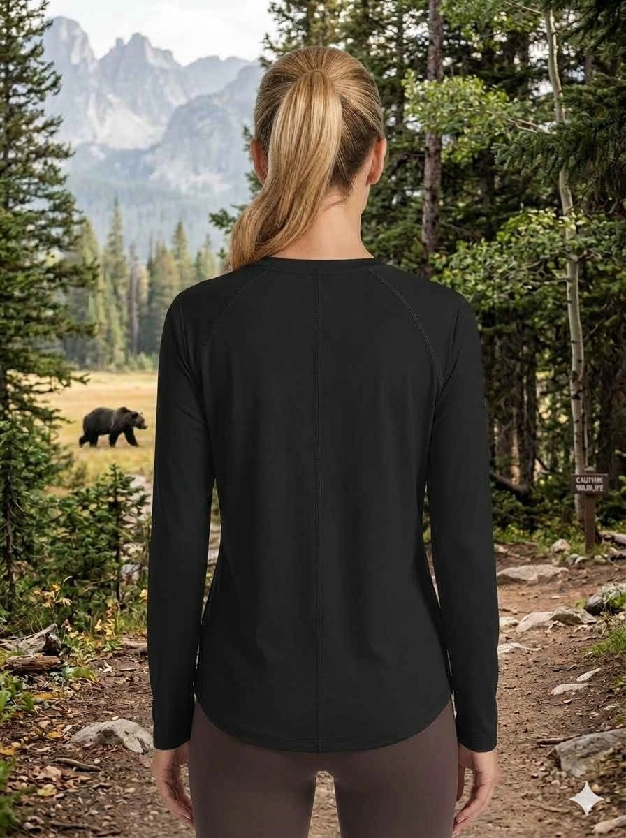 A woman viewed from behind wearing a black long-sleeve athletic top and brown leggings, standing on a forest trail with mountains and trees in the background.