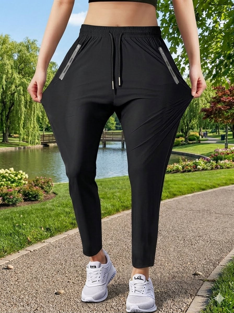 A person outdoors wearing black athletic jogger-style pants with side zip pockets and a drawstring waist, shown from the torso down on a park path near water.