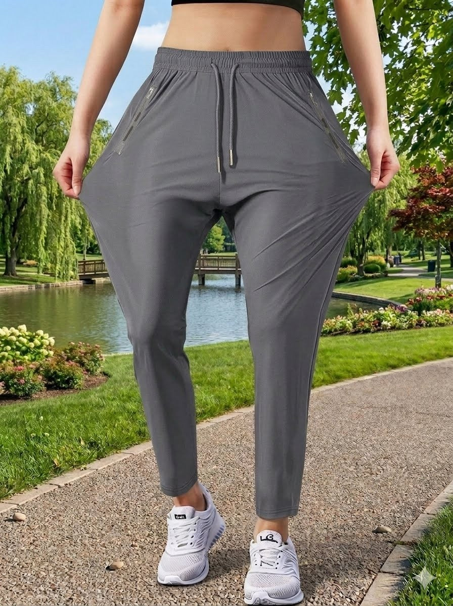 A person outdoors wearing loose gray jogger-style pants, holding the sides of the legs to show stretch. The background shows a park path, green grass, trees, and a small pond.