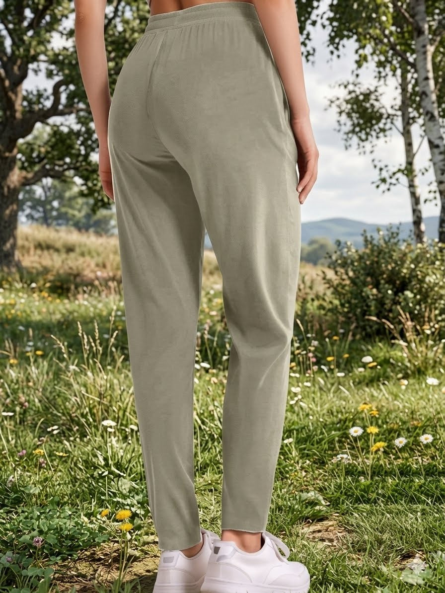 A person outdoors wearing light sage green jogger-style pants with white sneakers, photographed from the back in a grassy field with trees and hills in the background.