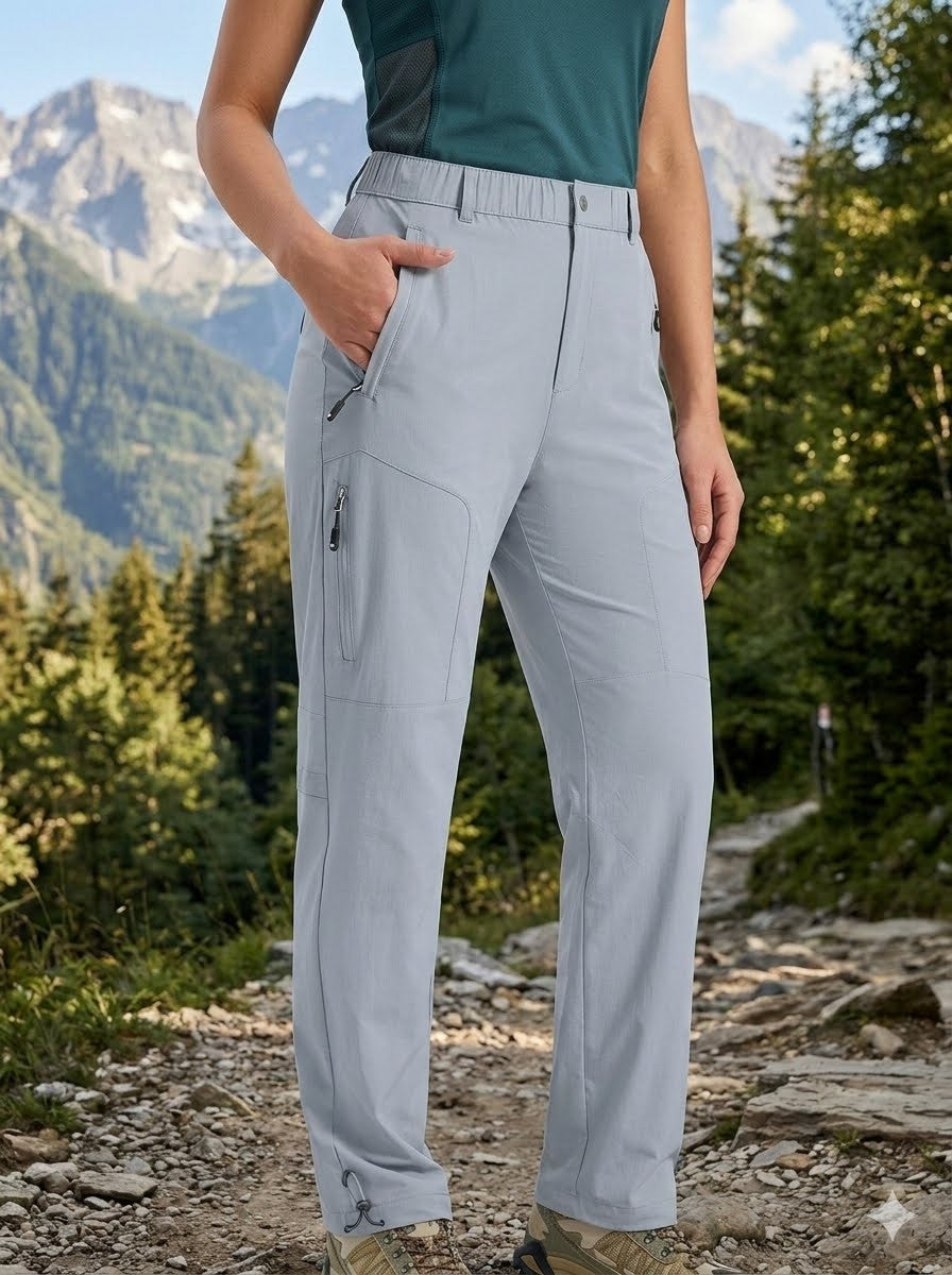 A person outdoors on a mountain trail wearing light gray hiking pants with zippered pockets and a dark green sleeveless top. Mountains and trees are visible in the background.