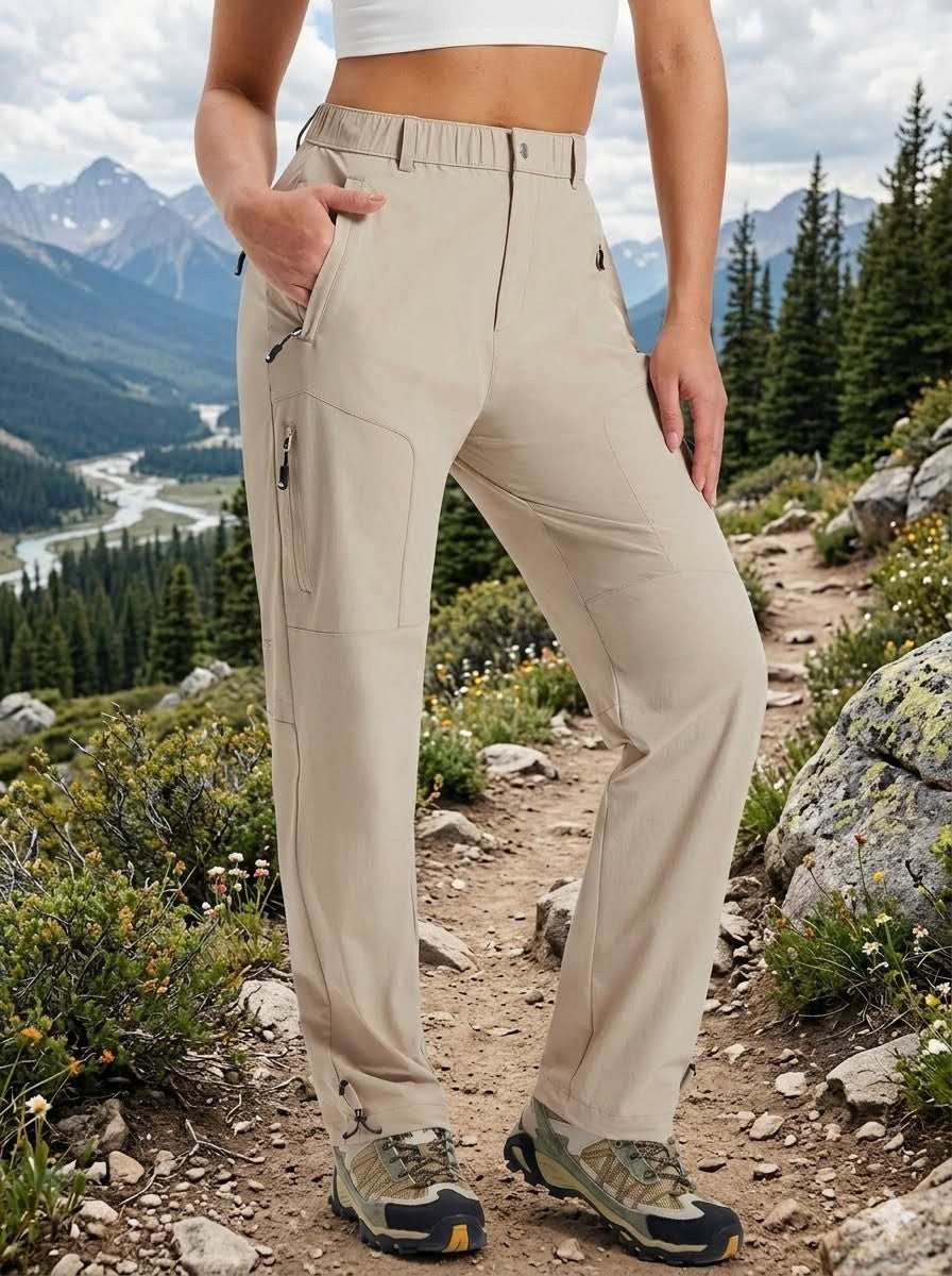 A person wearing beige hiking pants with zippered pockets stands on a rocky mountain trail with trees and mountains in the background.