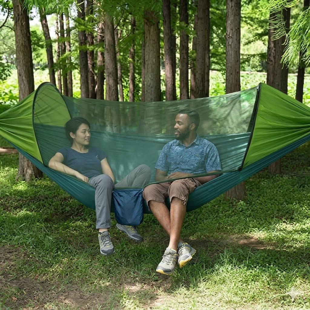 A man and a woman relax inside a green camping hammock with an attached mosquito net, suspended between trees in a grassy forest clearing. big green bear