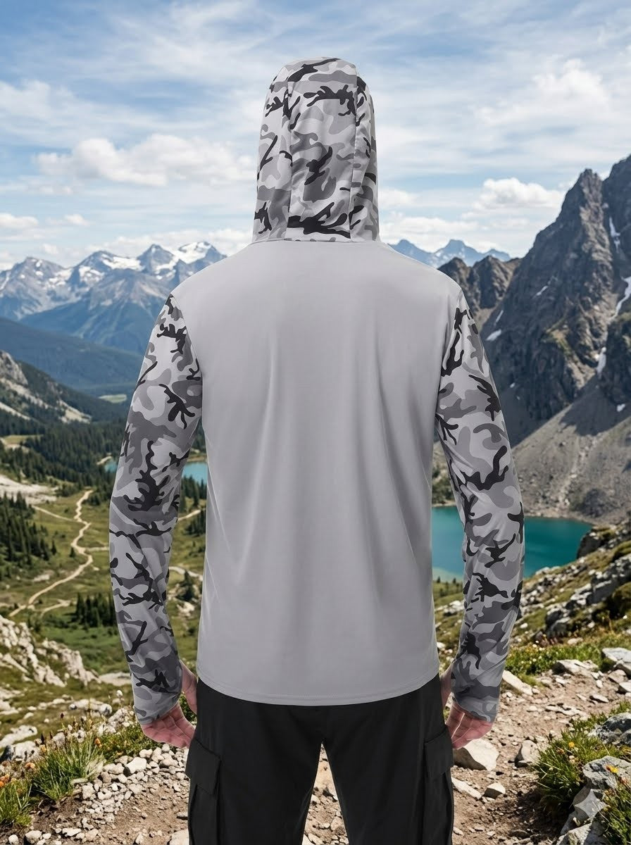 A person standing outdoors in a mountain landscape, viewed from the back, wearing a light gray hooded jacket with gray-and-black camouflage sleeves and hood, paired with dark pants.