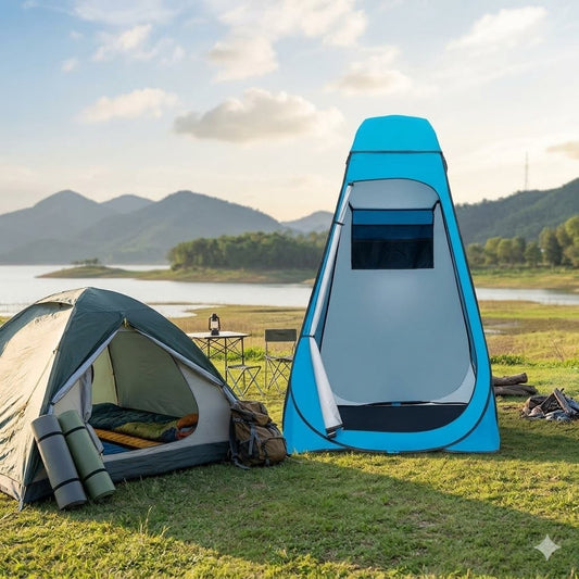 A bright blue pop-up shower tent stands open on grass beside a larger green and gray camping tent, with a lake, trees, and mountains in the background. big green bear