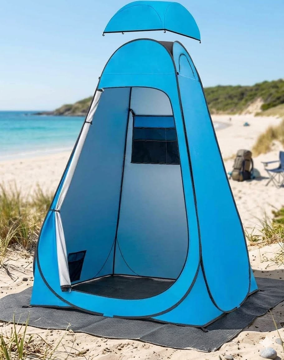 A bright blue pop-up beach tent with a matching rain hood on top, set up on a sandy beach with the ocean and dunes in the background.