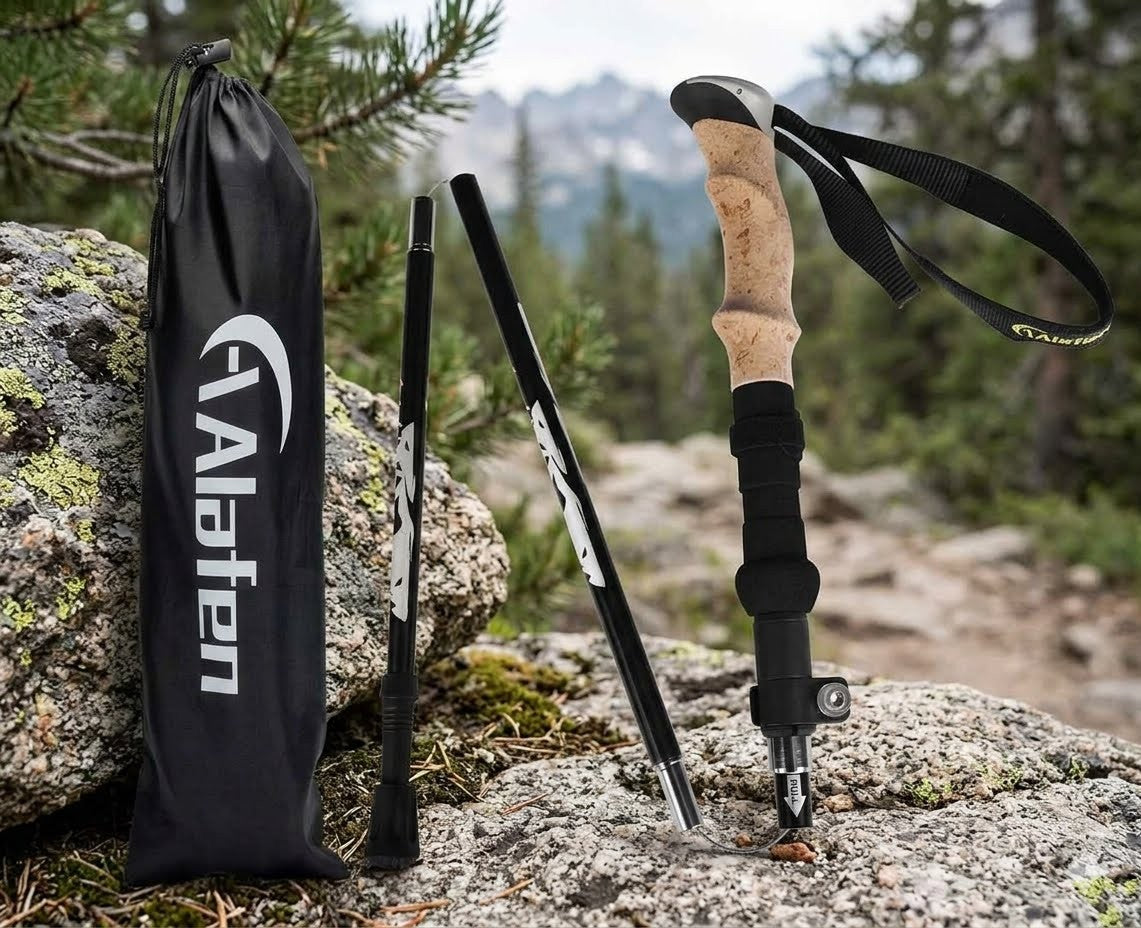 A set of outdoor hiking poles displayed on a rocky mountain path, including one fully extended pole with a cork and black handle, one folded pole, and a black drawstring carrying bag with the Alafan brand logo. The background shows a blurred forest and mountain landscape.