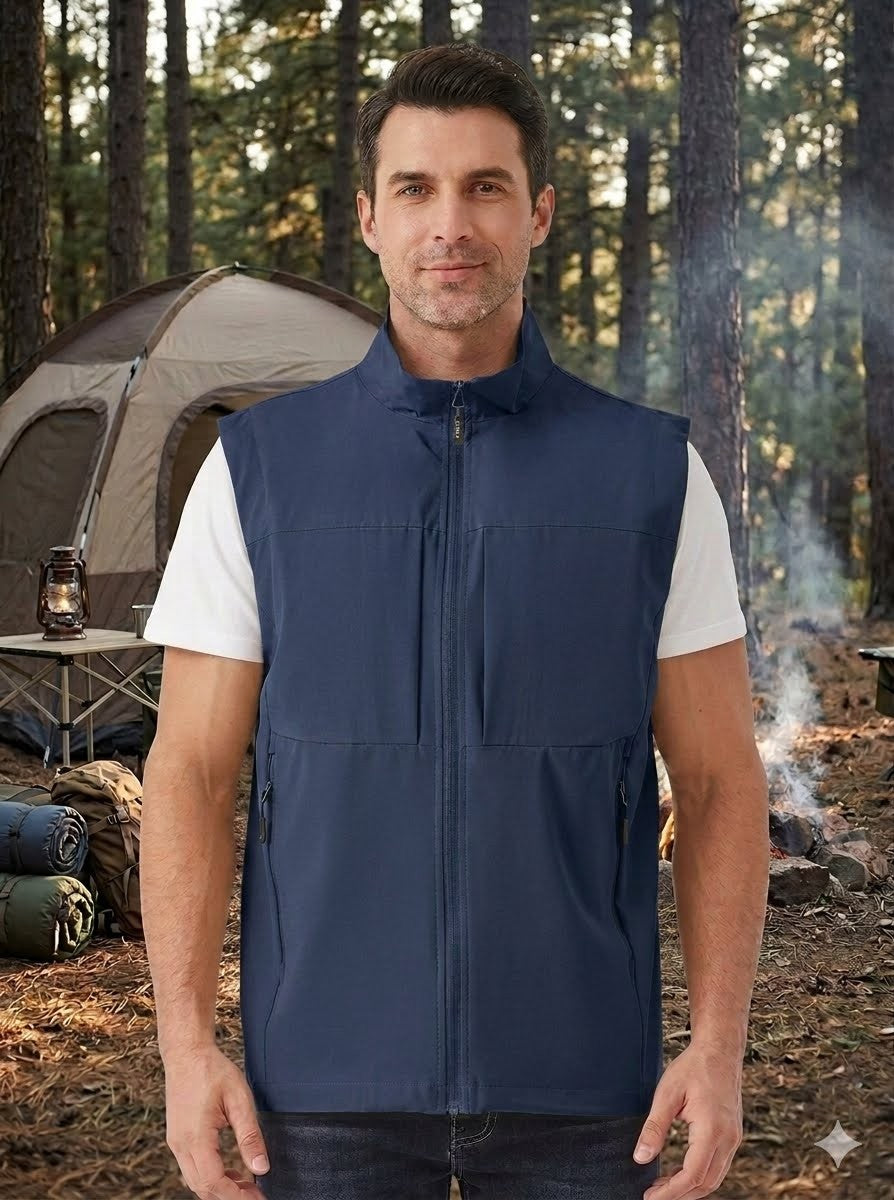 A man wearing a navy blue zip-up vest over a white t-shirt, standing in a forest campsite setting with a tent and a campfire in the background. big green bear biggreenbear.com