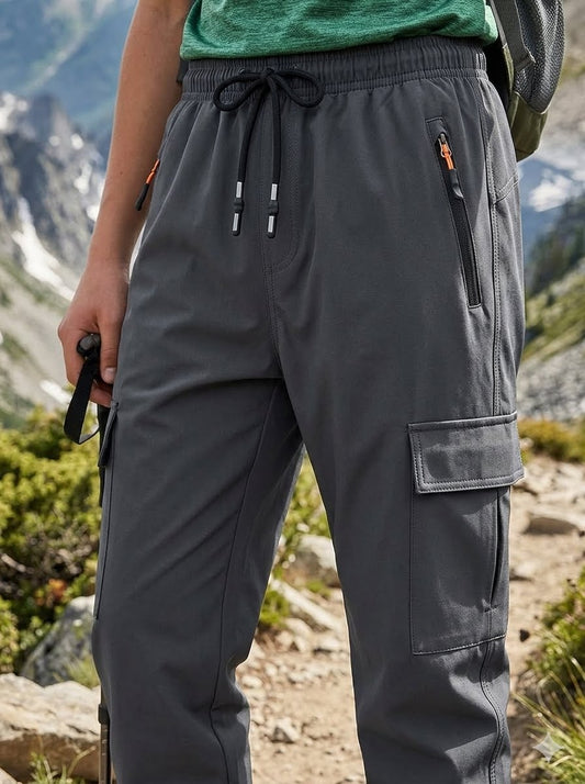 Dark gray outdoor cargo pants worn by a hiker in a mountain setting, featuring an elastic drawstring waist, zippered pockets, and a large cargo pocket on the thigh. big green bear biggreenbear.com