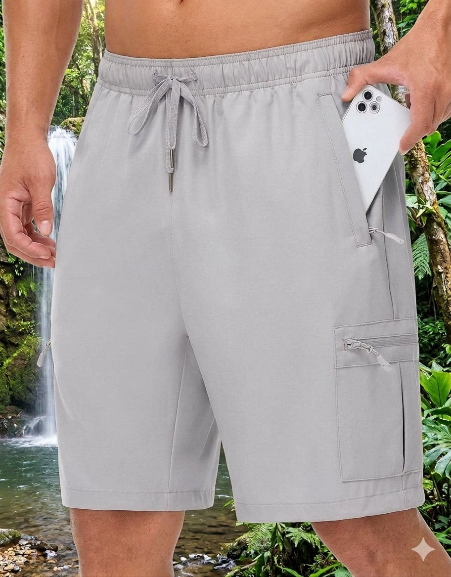 A man wearing light gray drawstring shorts with cargo pockets, standing in front of a lush tropical waterfall background. A white smartphone is tucked into the side pocket.