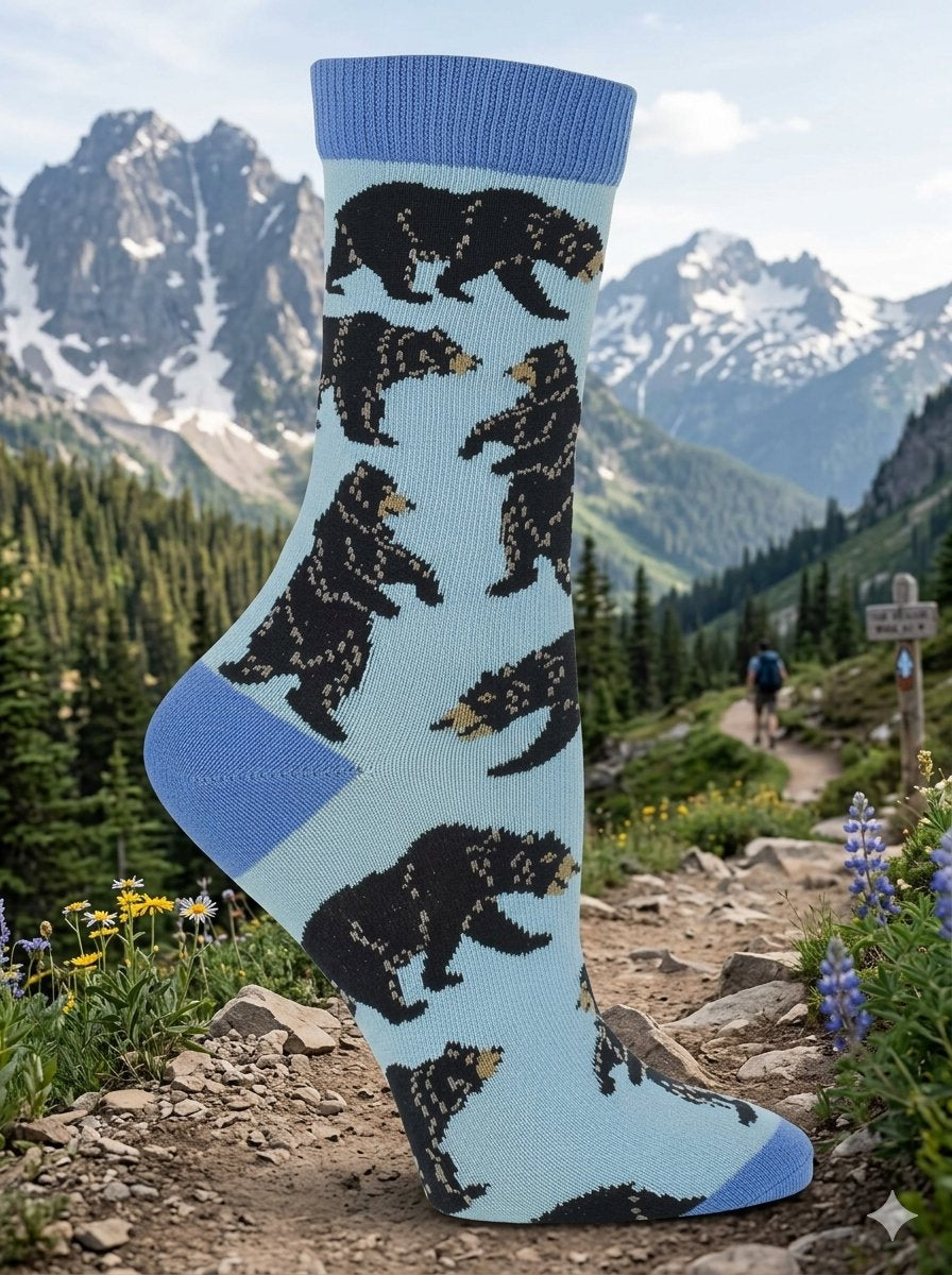 A single light blue crew sock with a pattern of black bears and small tan details, displayed on a rocky mountain trail with a hiker and snowy peaks in the background.